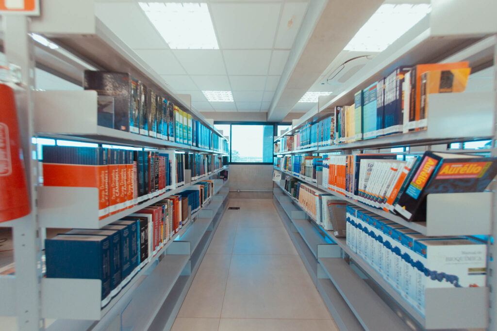 Corredor da Biblioteca da Faculdade Zarns Itumbiara. Na foto duas estantes, uma a esquerda e outra a direita com 3 prateleiras. No fundo, uma luminosidade vindo de uma janela.